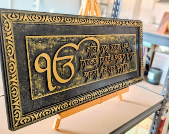 Op de afbeelding: Een rechthoekig wandbord met een zwarte en gouden verweerde afwerking en een decoratieve rand. Het bord toont het gouden Gurmukhi-symbool Ik Onkar en de Mool Mantar-tekst in verhoogde gouden letters op een donkere, getextureerde achtergrond.