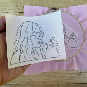 May include: A black and white line drawing of a person wearing sunglasses and smoking a cigarette. The drawing is on a piece of white fabric, held in a hand. A pink fabric and embroidery hoop are in the background.