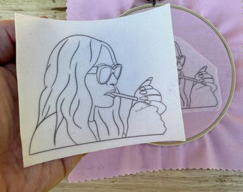 Puede incluir: Un dibujo lineal en blanco y negro de una persona con gafas de sol fumando un cigarrillo. El dibujo está sobre un trozo de tela blanca, sostenido en una mano. Una tela rosa y un bastidor de bordado están en el fondo.