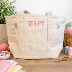 May include: Beige canvas tote bag with a front pocket and embroidered details. Features the letters "NDLPT" in pink, along with a spool of thread, a needle, and scissors. Perfect for carrying sewing supplies.
