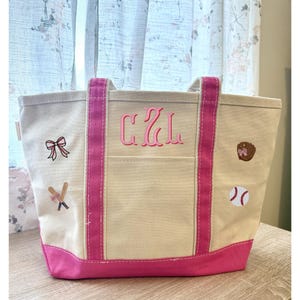 May include: A beige canvas tote bag with pink accents, including the bottom, handles, and side stripes. The bag features a pink monogram and embroidered baseball-themed patches, such as a baseball, glove, and crossed bats.