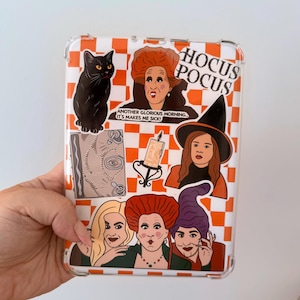 May include: Clear phone case with a white and orange chequered pattern, featuring stickers of a black cat, a spell book, a candle, and three witches. The text "Hocus Pocus" is visible.