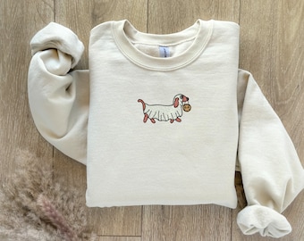 Könnte beinhalten: Ein cremefarbener Pullover mit einem Cartoon-Bild eines Dackels, der als Geist verkleidet ist und einen Kürbis hält. Der Pullover hat einen Rundhalsausschnitt und lange Ärmel.