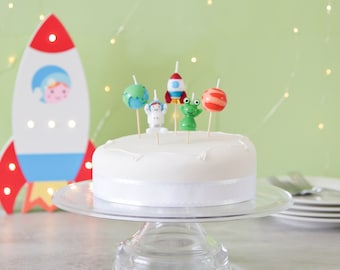 May include: A white cake with a white ribbon on a glass cake stand. The cake is decorated with astronaut, rocket, planet, and alien candles.
