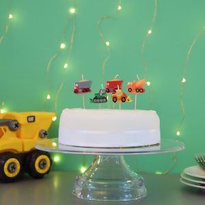Könnte beinhalten: Ein weißer Kuchen mit Baufahrzeug-Kerzen darauf. Der Kuchen steht auf einem gläsernen Kuchenständer. Im Vordergrund befindet sich ein gelber Spielzeug-Kipplaster.