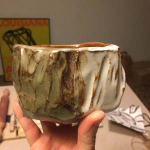 Puede incluir: Una taza pequeña de cerámica hecha a mano con un exterior rugoso y texturizado. Un lado está esmaltado en blanco, mientras que el otro lado es verde y marrón moteado.