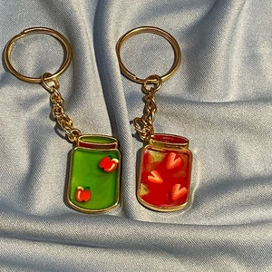 Op de afbeelding: Twee gouden sleutelhangers met bedels in de vorm van potten. Een bedel is groen met rode harten erin, de andere is rood met witte harten erin.