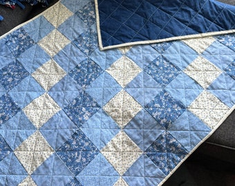 May include: A blue and white patchwork quilt with a diamond pattern. The quilt features various shades of blue and white fabrics, including floral and solid patterns. The back of the quilt is a solid navy blue with a diamond stitch pattern.
