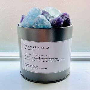 以下が含まれることがあります： 三日月マークと「manifest」の文字が書かれた白いラベルが付いた銀色の金属製容器。ラベルには、「ask, believe, receive. mantra: I am the vibration of my dreams. ethical | eco-friendly | empowering. Ethereal Haze Co. Aromatherapy Crystal Diffuser」というテキストも記載されています。容器は青と紫のクリスタルで満たされています。