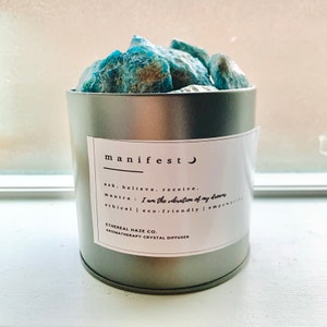 Puede incluir: Un recipiente de metal plateado con una etiqueta blanca que dice "manifest" y un símbolo de luna creciente. La etiqueta también incluye el texto "ask. believe. receive." y "mantra: I am the vibration of my dreams." El recipiente está lleno de cristales azul verdosos. La etiqueta también incluye el texto "ETHEREAL HAZE CO. AROMATHERAPY CRYSTAL DIFFUSER."