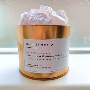 Puede incluir: Un recipiente de metal dorado con una etiqueta blanca que dice "manifest" y un pequeño símbolo de luna creciente. La etiqueta también incluye el texto "ask. believe. receive." y "mantra: I am the vibration of my dreams". El recipiente está lleno de cristales de cuarzo rosa. La etiqueta también incluye el texto "ETHEREAL HAZE CO. AROMATHERAPY CRYSTAL DIFFUSER".