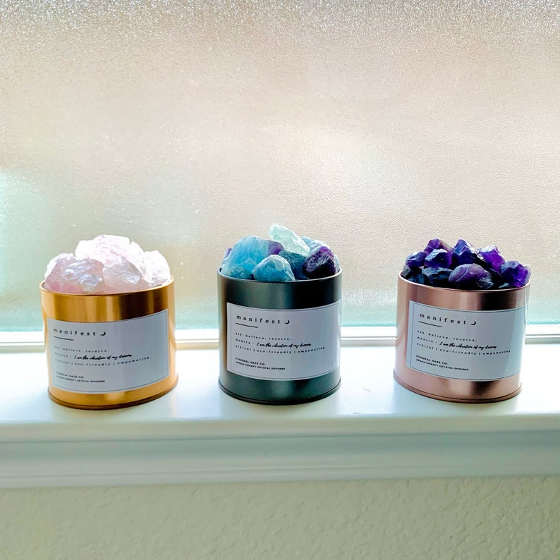 May include: Three small metal containers filled with crystals sit on a windowsill. The containers are gold, gray, and rose gold. Each has a label with the word "manifest" and other text. The containers hold pink, blue, and purple crystals.