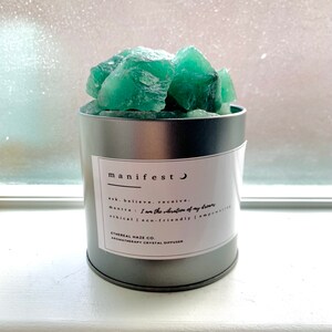 Puede incluir: Un difusor de cristal verde en un recipiente de metal plateado. El recipiente tiene una etiqueta que dice "manifest" y "I am the vibration of my dreams".