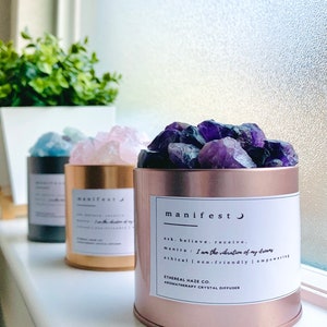 May include: Three aromatherapy crystal diffusers in a row. The front diffuser is rose gold and filled with purple crystals. The label reads "manifest" and includes the mantra "I am the vibration of my dreams". The other diffusers are gold and black.