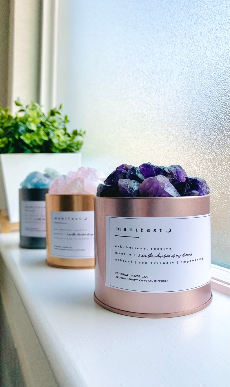 May include: Three aromatherapy crystal diffusers in a row. The front diffuser is rose gold and filled with purple crystals. The label reads "manifest" and includes the mantra "I am the vibration of my dreams". The other diffusers are gold and black.