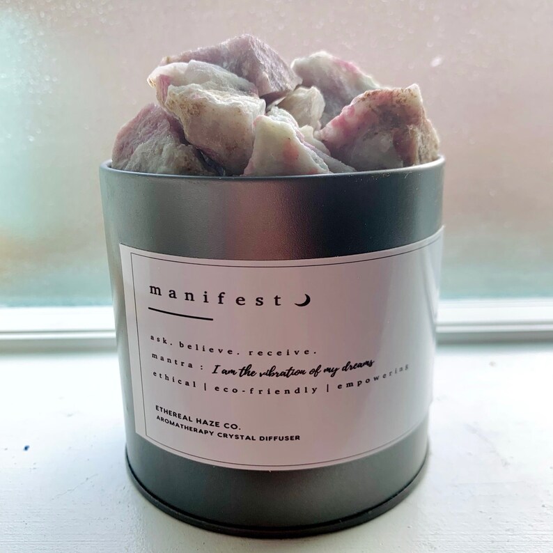 May include: A dark gray aromatherapy crystal diffuser filled with rough, pink and white crystals. The diffuser has a white label with the word "manifest" and other text. The text includes "ask, believe, receive" and "I am the vibration of my dreams."