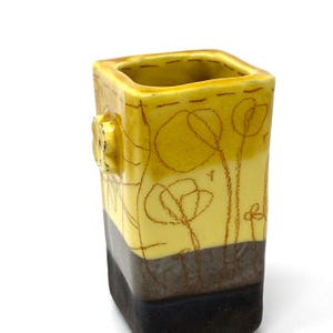 May include: A square ceramic vase with a yellow glaze and brown accents. The vase has a simple design with a flower pattern and a small button-like detail on the side.