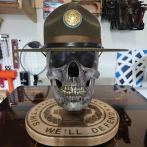 May include: A detailed skull sculpture wearing a green and brown military hat with a gold emblem. The skull rests on a wooden base engraved with the words "THIS WE'LL DEFEND". The teeth are gold-colored.