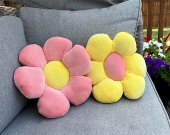 May include: Two plush flower-shaped pillows in pink and yellow. The pillows are sitting on a gray couch.