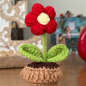 以下が含まれることがあります： 赤い花と緑の葉が付いた、かぎ針編みで作った植木鉢。鉢は茶色で、濃い茶色の縁取りがある。