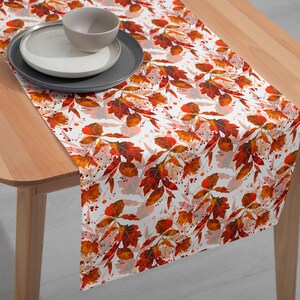 May include: A white table runner with a fall leaf pattern in shades of orange and red. The runner is on a wooden table with a bowl and plate on top.