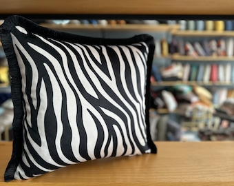 Puede incluir: Cojín decorativo de color blanco y negro con estampado de cebra y ribete de flecos negros.