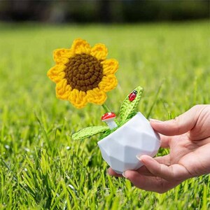 May include: A crocheted sunflower with a brown center and yellow petals in a white pot with a green leaf and a red ladybug. The pot is made of a textured material and has a geometric design.