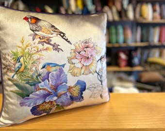 May include: A decorative throw pillow with a beige background featuring a floral design with a pink flower, purple irises, and three birds. The birds are a brown finch, a blue bird, and a blue and yellow bird.