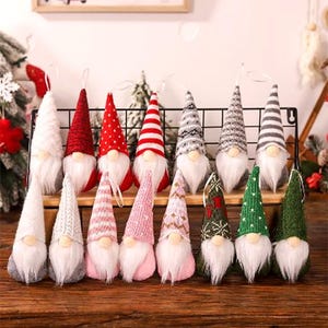 Può includere: Una collezione di ornamenti natalizi a forma di gnomi, con barbe bianche, cappelli a forma di cono e nasi rotondi. I cappelli sono di vari colori e motivi, tra cui rosso, bianco, grigio, rosa e verde. Ogni gnomo ha un anello per appenderlo.