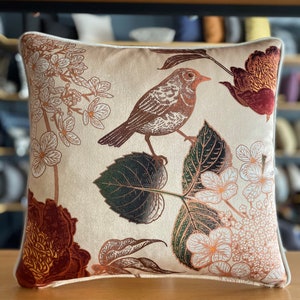 May include: A cream-colored throw pillow with a floral and bird design. The pillow features a brown bird with a red breast, green leaves, and white flowers with orange accents.