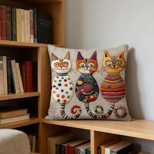 Könnte beinhalten: Ein quadratisches Zierkissen mit hellgrauem Stoffbezug und einer Stickerei von drei Katzen. Die linke Katze ist weiß mit bunten Punkten, die mittlere Katze hat einen gemusterten Körper und die rechte Katze ist mit horizontalen Streifen versehen.