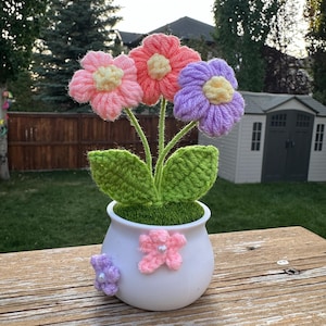 Op de afbeelding: Een gehaakt bloemstuk in een witte pot met groene bladeren. De bloemen zijn roze, lichtroze en paars.