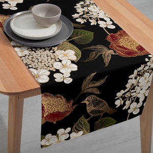 Può includere: Runner da tavolo nero con un design floreale con fiori bianchi, dorati e bordeaux e un'illustrazione di uccelli. Il runner è posto su un tavolo di legno, con piatti e una ciotola sopra.