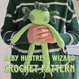 May include: A light green crochet doll with large eyes and long limbs is held in front of a green and white striped sweater. The doll has a whimsical design with a crown-like feature on its head. The text "BABY HUNTRESS WIZARD CROCHET PATTERN" is displayed below.