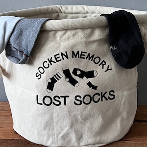 Könnte beinhalten: Ein weißer Canvas-Wäschekorb mit schwarzem Text, der "Sock Memory Lost Socks" lautet. Der Korb hat eine schwarz-weiße Illustration von Socken darauf. Es befinden sich Socken im Korb.