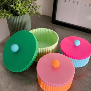 Könnte beinhalten: Drei runde, gerippte Aufbewahrungsbehälter mit Deckeln. Ein Behälter ist hellgrün mit einem dunkelgrünen Deckel und blauem Knopf. Ein weiterer ist rosa mit einem orangefarbenen Knopf. Der dritte ist blau mit einem rosafarbenen Deckel und blauem Knopf.