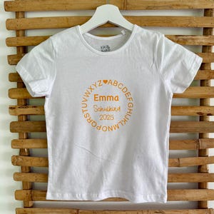 Könnte beinhalten: Weißes T-Shirt mit einem orangefarbenen runden Design. Das Design enthält den Namen "Emma", das Wort "Schulkind" und das Jahr "2025". Der Text wird von Buchstaben und Herzen umkreist. Das T-Shirt hängt an einem weißen Kleiderbügel vor einem Bambushintergrund.