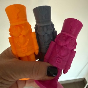 Könnte beinhalten: Drei Nussknacker-Figuren in leuchtendem Orange, Dunkelgrau und kräftigem Pink. Jede Figur hat einen hohen Hut, eine Uniform mit Gürtel und einen runden Sockel. Die Nussknacker sind mit detaillierten Gesichtszügen, einschließlich Schnurrbart und Bart, geformt.