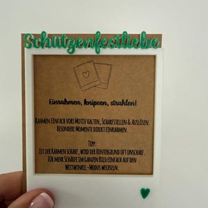Könnte beinhalten: Ein weißer rechteckiger Rahmen mit dem grünen Schriftzug "Schützenfestliebe" oben und einem kleinen grünen Herz unten. Der Rahmen enthält eine braune Karte mit dem schwarzen Text "Einrahmen, knipsen, strahlen!" und Anweisungen zur Fotografie.