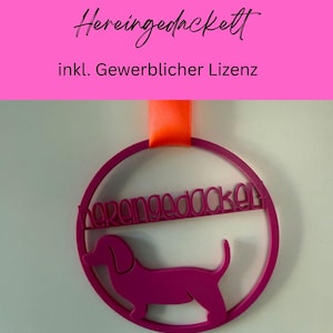 Könnte beinhalten: Ein magentafarbenes Dackel-Ornament mit dem Text "Hereingedackelt" in einem kreisförmigen Rahmen, aufgehängt an einem orangefarbenen Band. Der obere Teil des Bildes enthält Text auf Deutsch.