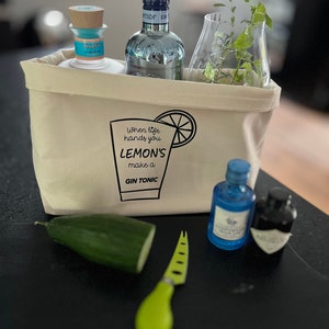 Könnte beinhalten: Eine weiße Stofftasche mit dem Text "When life hands you lemons make a gin tonic" und einer Zitronenscheibengrafik. Die Tasche enthält zwei Flaschen Gin, eine Glasvase mit frischer Minze, eine Gurke und ein grünes Käsemesser.