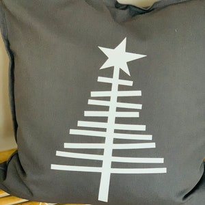Könnte beinhalten: Ein graues Kissen mit einem weißen geometrischen Weihnachtsbaumdesign. Der Baum hat einen Stern oben und besteht aus horizontalen Linien.