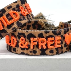 Könnte beinhalten: Zwei Armbänder mit Leopardenmuster und orangefarbenem, gesticktem Text "WILD & FREE". Die Armbänder haben einen beigefarbenen Hintergrund mit schwarzen Flecken und einem Herzsymbol. Quasten sind an einem Armband befestigt.