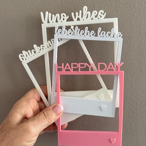 Könnte beinhalten: Eine Hand hält mehrere weiße und rosa Fotorahmen-Ausschnitte. Die Rahmen tragen die Texte "Vino vibes", "lebe liebe lache", "Glucksmo" und "HAPPY DAY". Einige Rahmen haben Herzdetails.