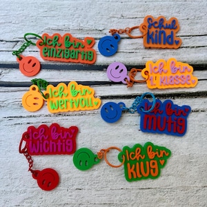 Könnte beinhalten: Eine Sammlung bunter Schlüsselanhänger mit Smiley-Gesichtern und deutschem Text. Die Schlüsselanhänger sind in verschiedenen Farben erhältlich, darunter Orange, Grün, Pink und Blau. Jeder Schlüsselanhänger hat einen anderen Spruch, wie "Ich bin einzigartig" und "Schul Kind".