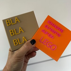 Könnte beinhalten: Zwei quadratische Untersetzer werden in einer Hand gehalten. Einer ist beige mit dem Wort "BLA" dreimal in Gelb. Der andere ist orange mit den Worten "ICH HORRE DU TRITTS AUFEN LEGO" in Pink.