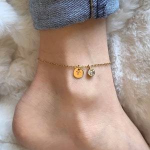 May include: A gold anklet featuring two charms: a circular disc with the Aries symbol and a clear, round gemstone. The anklet is worn on an ankle, with a denim cuff visible in the background. The background is a fluffy, white material.