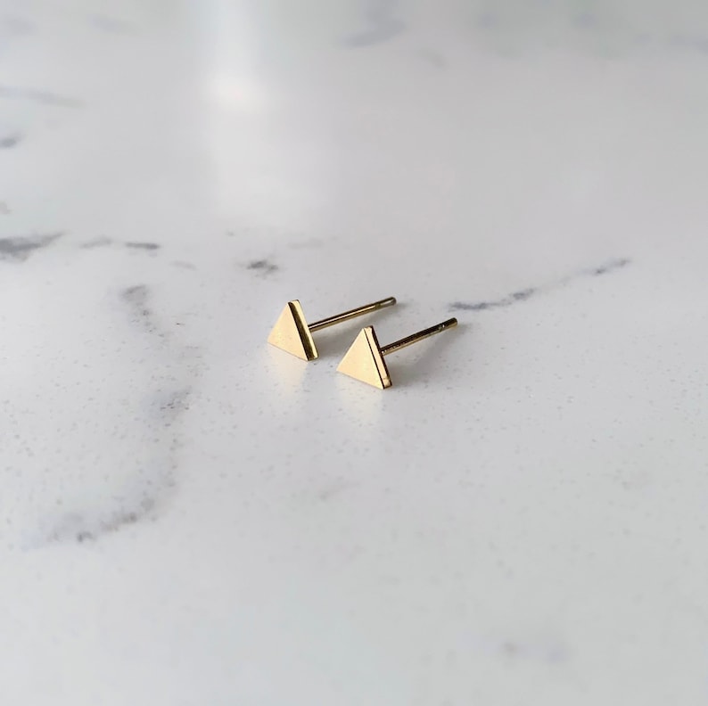 May include: A pair of gold triangle stud earrings. The earrings are small and simple, making them a versatile accessory for any occasion.