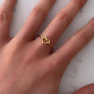 May include: A delicate gold ring with a heart-shaped design. The ring is made from thin, shiny gold metal and is worn on a finger. The heart is formed by a continuous line, creating an open, airy look. A simple and elegant piece of jewellery.