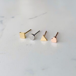 May include: Four gold stud earrings with different shapes: a square, a circle, a triangle, and a heart. The earrings are on a white marble surface.